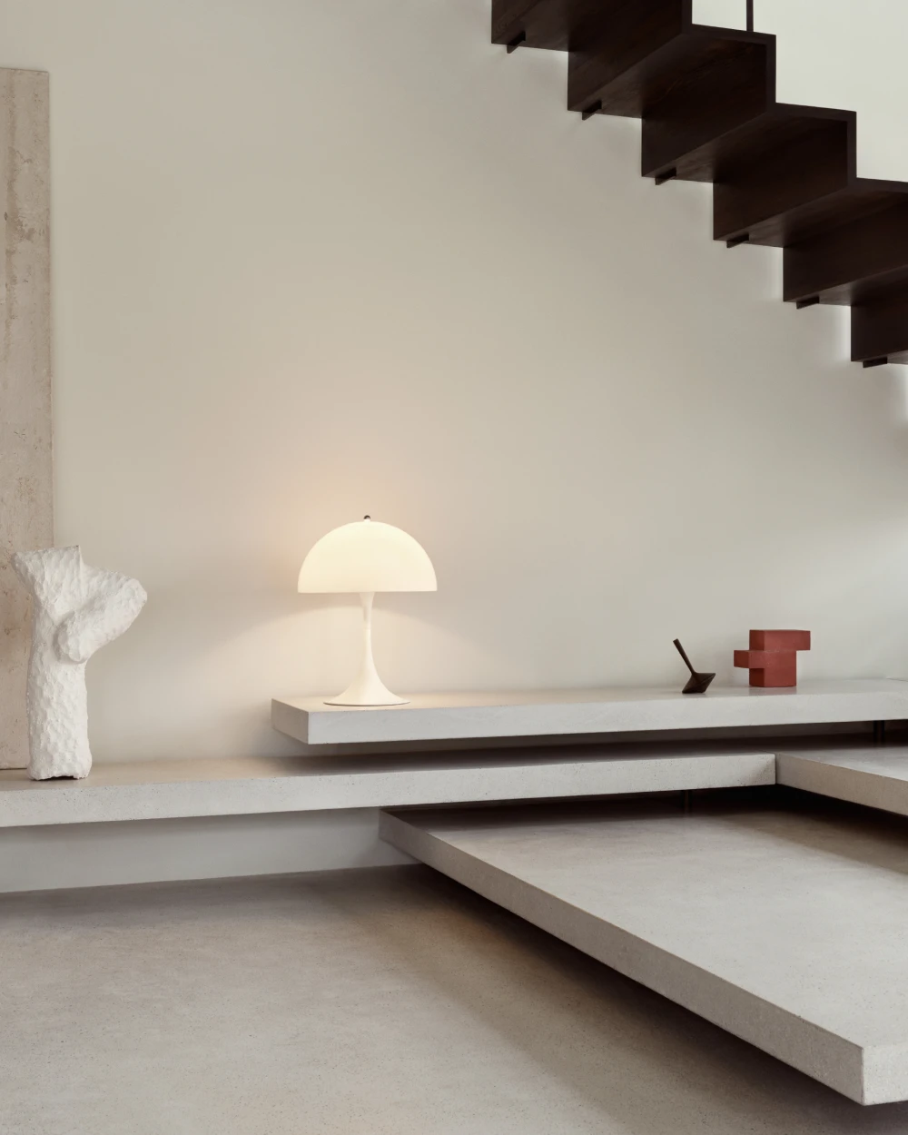 A white Panthella 400 Table Lamp glows on a floating shelf in a minimalist room with concrete surfaces. © 2026 Louis Poulsen.