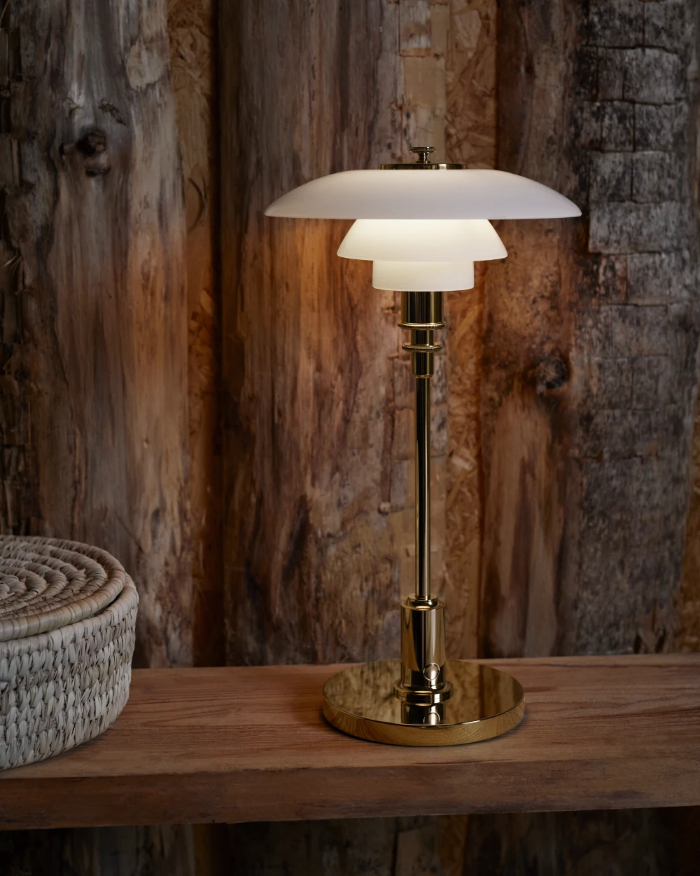 La lampe portable PH 2/1, finition laiton et abat-jour en verre opalin blanc, éclaire une table en bois devant un mur rustique. © 2026 Louis Poulsen.