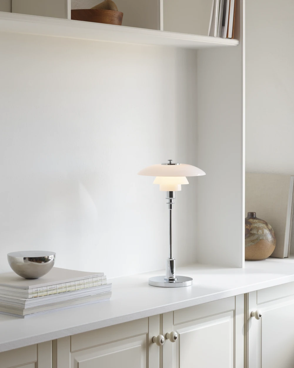 La lampe portable PH 2/1, avec ses abat-jour en verre blanc et son socle chromé, est posée sur un buffet blanc. © 2026 Louis Poulsen.