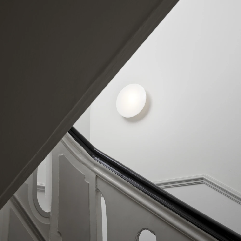 A white AJ Eklipta Wall Lamp glows on the wall of a stairwell with a classic banister. © 2026 Louis Poulsen.