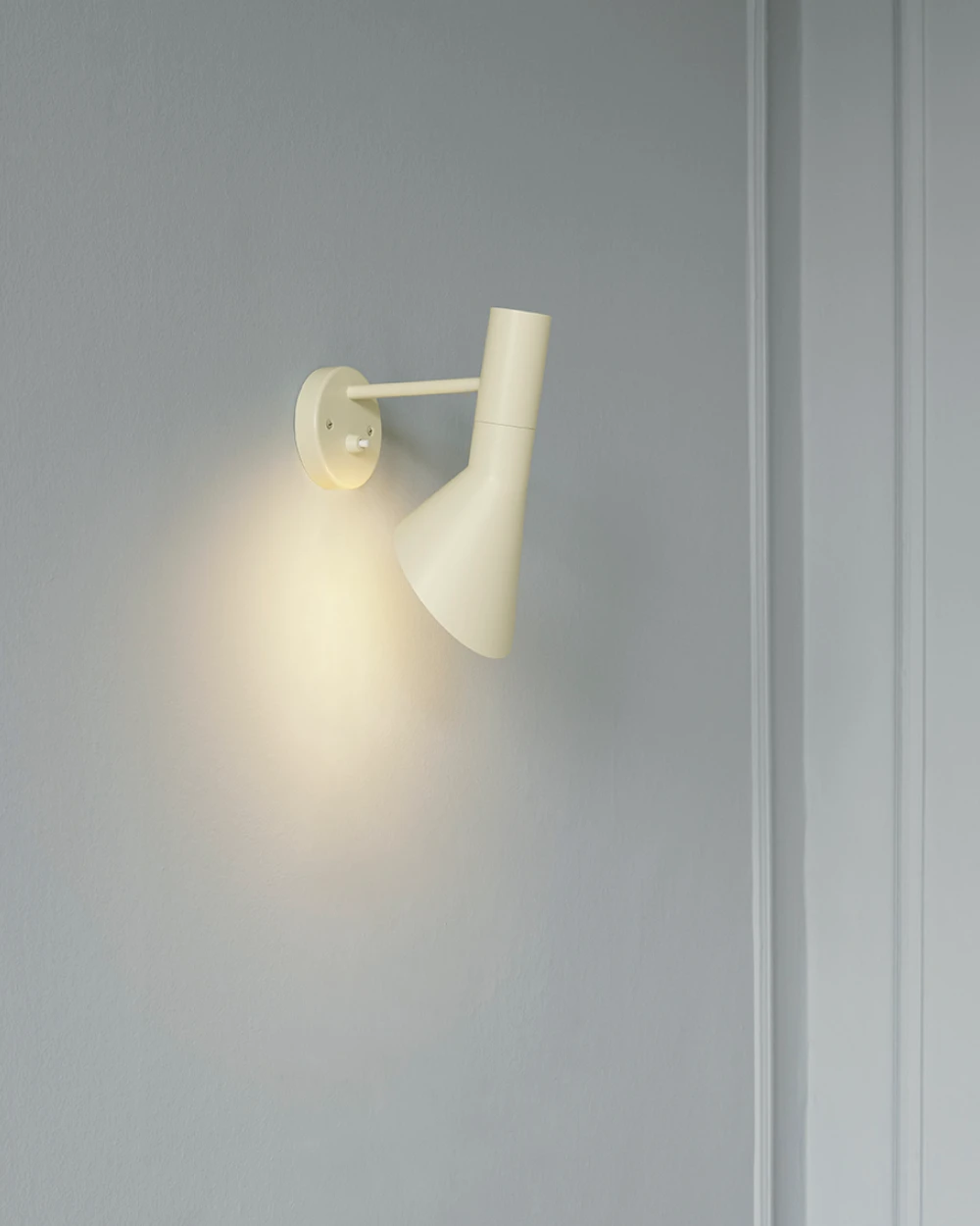 A white AJ Wall Lamp casts a soft cone of light on a grey wall. © 2026 Louis Poulsen.