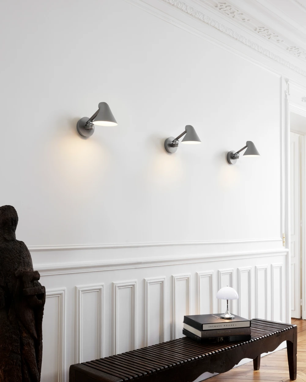 Three grey NJP Wall Lamp wall lights illuminate the wall above a bench, with a white Panthella 160 Portable Lamp on books. © 2026 Louis Poulsen.