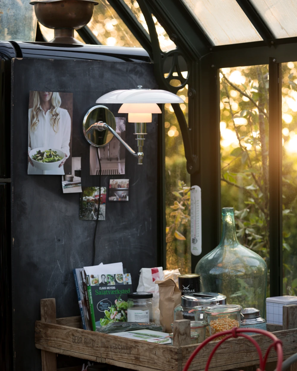 A PH 3/2 Wall Lamp with white shades glows in a greenhouse beside a dark wall and garden windows. © 2026 Louis Poulsen.