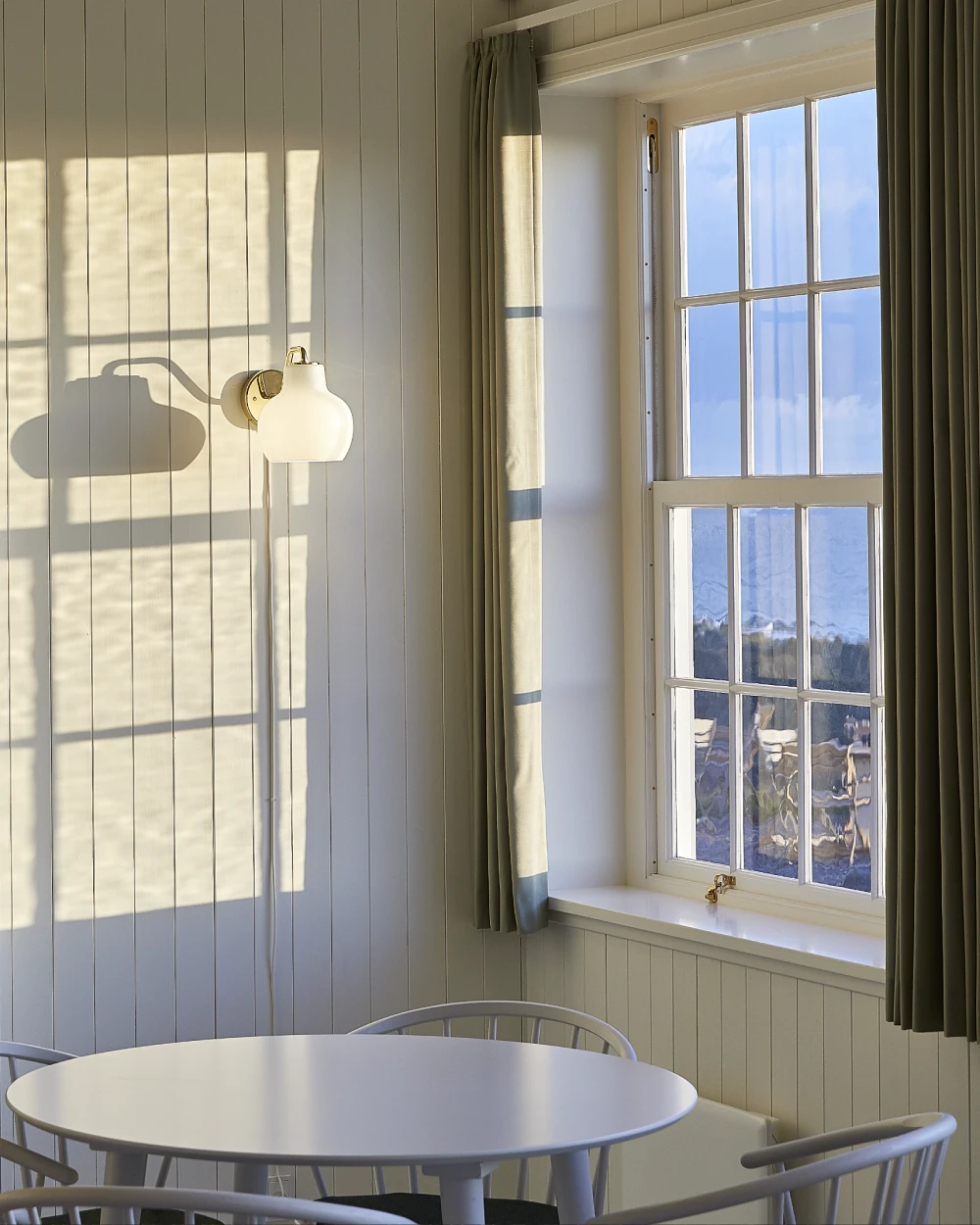 A VL Ring Crown 1 Wall Lamp with an opal glass shade on a light wall beside a window and round table. © 2026 Louis Poulsen.