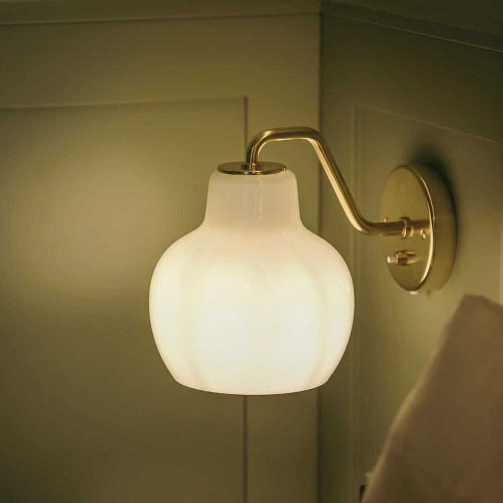 En VL Ring Crown 1 vägglampa med opalvitt glas och mässingsfinish sprider ett varmt sken mot en grön vägg. © 2026 Louis Poulsen.