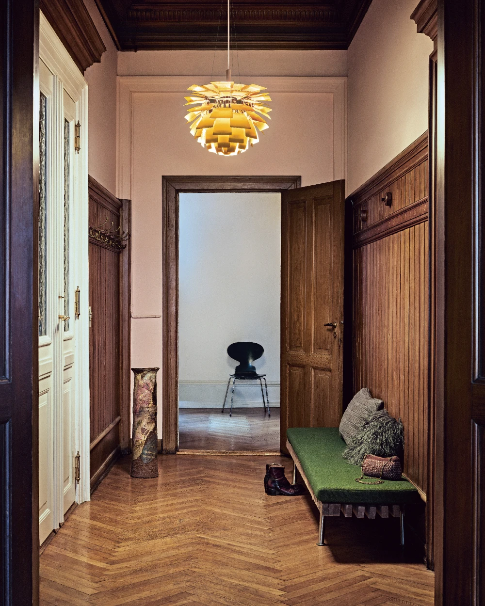 A PH Artichoke pendant casts a warm glow in a hallway with dark wood panelling, herringbone flooring and a green bench. © 2026 Louis Poulsen.