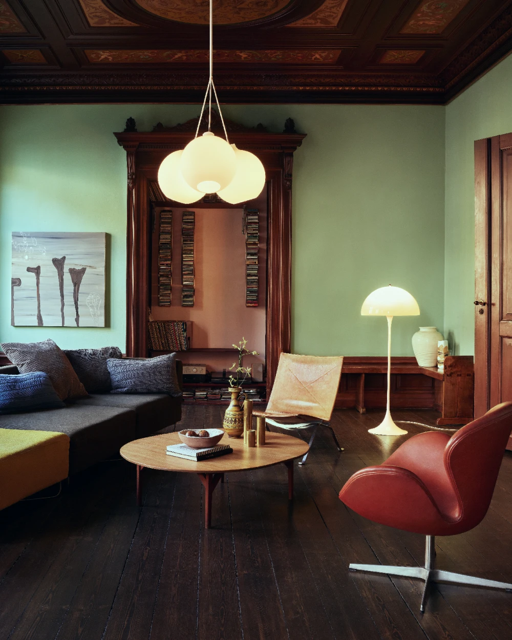 A VL Ring Crown 3-5-7 Pendant and a Panthella Floor Lamp light a living room with green walls and dark wood. © 2026 Louis Poulsen.