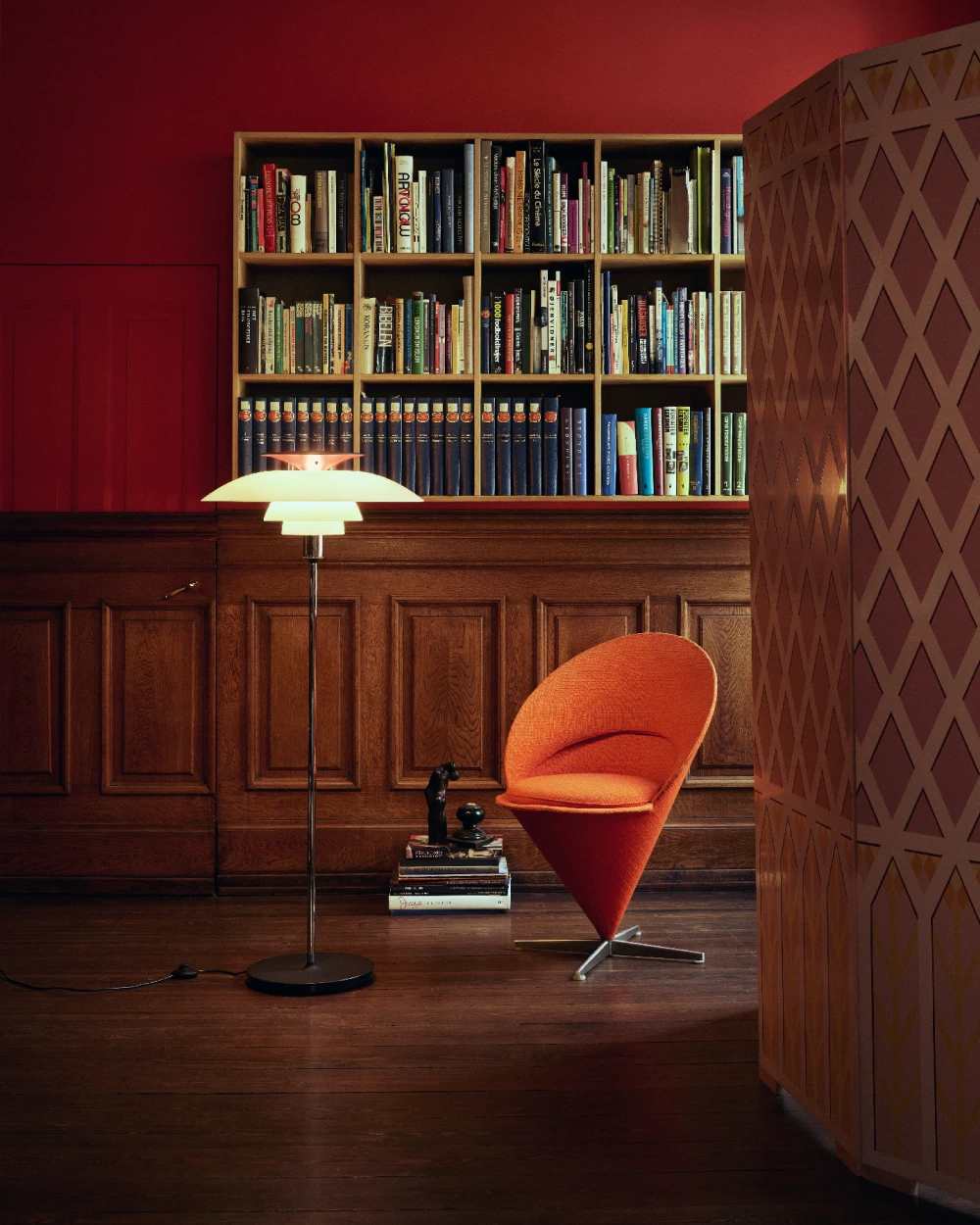 A PH 80 Floor Lamp with white shades illuminates a library with red walls, wood panelling and an orange chair. © 2026 Louis Poulsen.