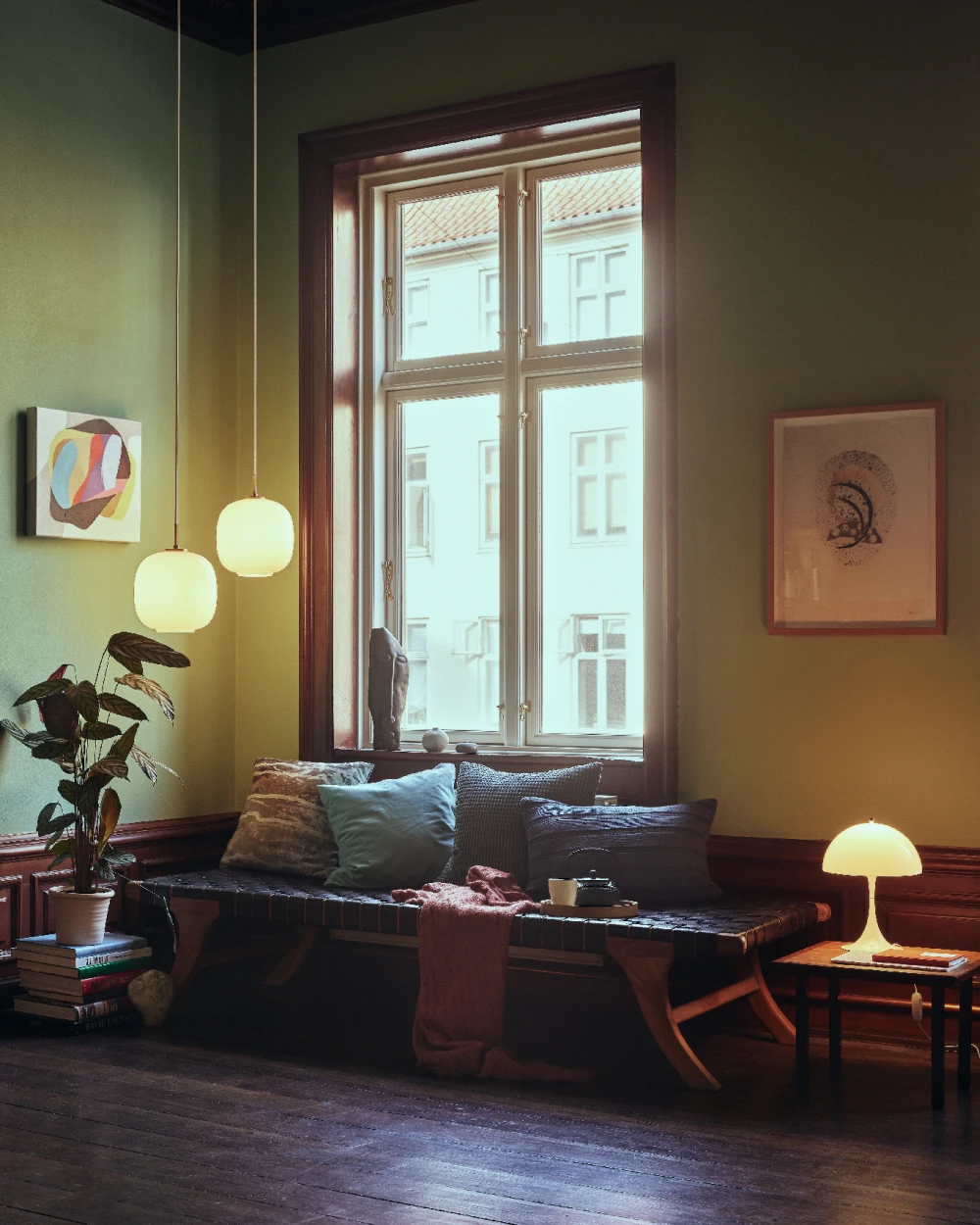 Two VL 45 Radiohus Pendants and a Panthella 250 Table Lamp glow in a green living room beside a window seat. © 2026 Louis Poulsen.