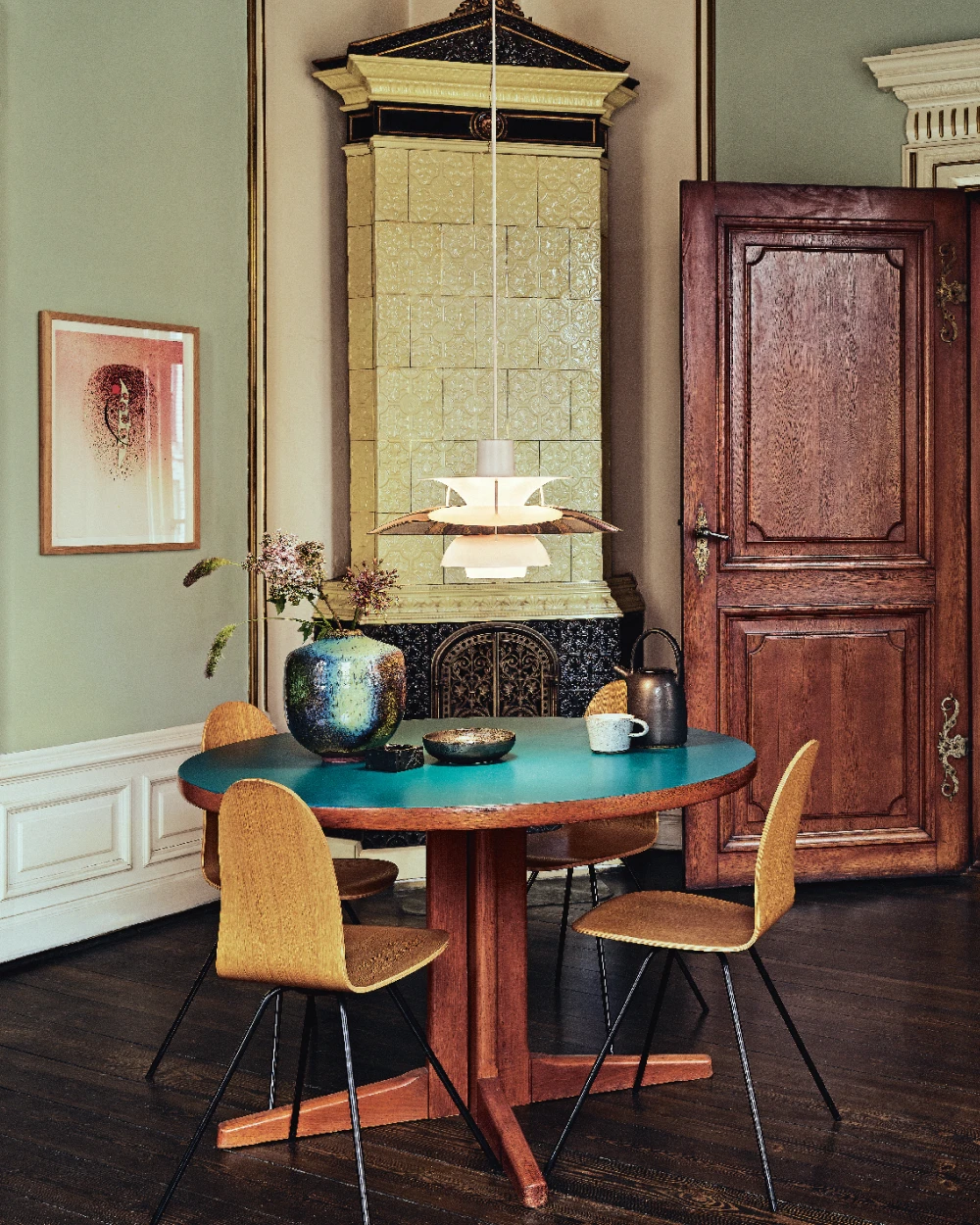A white PH 5 pendant lights a turquoise dining table in a colourful room with a tiled stove. © 2026 Louis Poulsen.