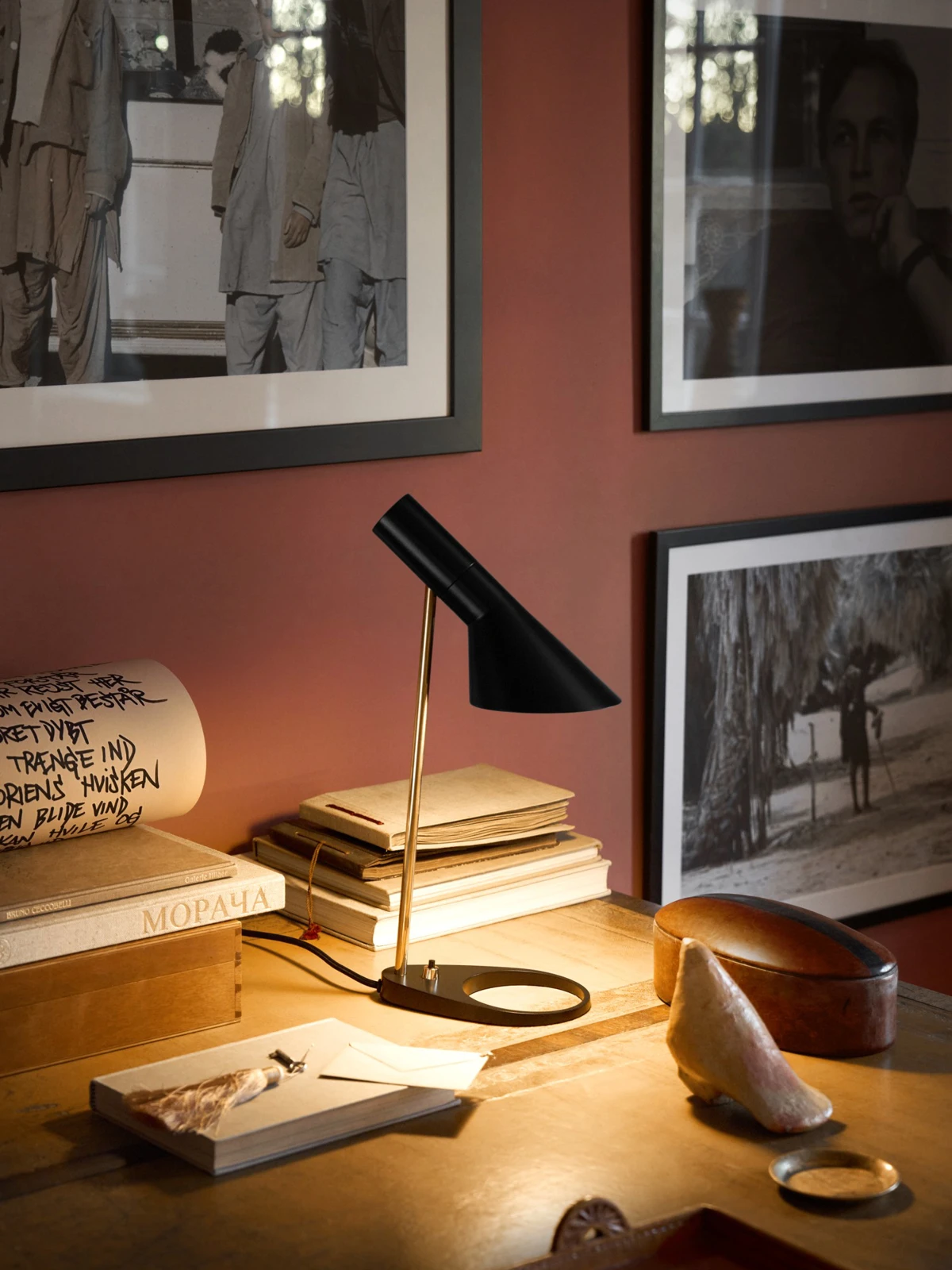 A black AJ Table Lamp with a brass stem lights a wooden desk beneath framed photographs. © 2026 Louis Poulsen.