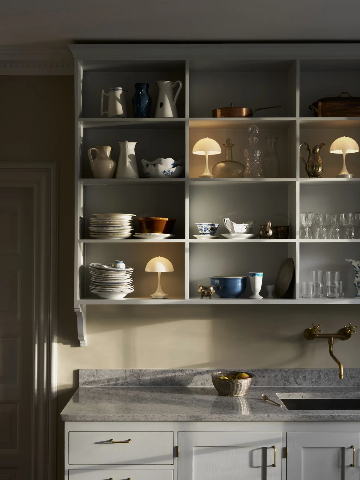 Three Panthella 160 Portable Lamps cast a warm glow among crockery on the shelves of a kitchen. © 2026 Louis Poulsen.