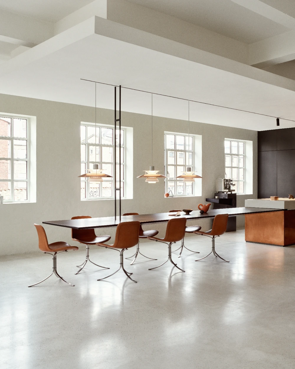 Kolme kuparinväristä PH 3½-3 Copper Pendant -riippuvalaisinta riippuu pitkän ruokapöydän yllä valoisassa, modernissa keittiössä. © 2026 Louis Poulsen.