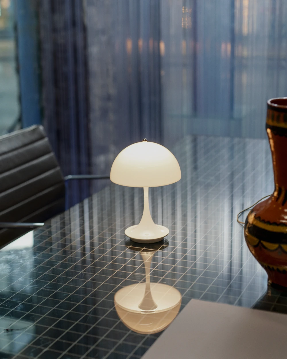 Une Panthella 250 Portable Lamp blanche éclaire une table en verre sombre quadrillée près de la fenêtre. © 2026 Louis Poulsen.