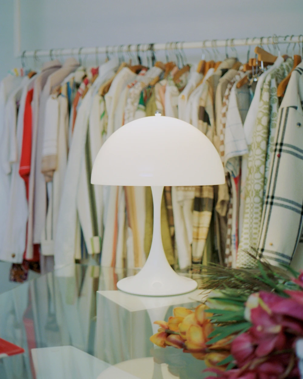 A white Panthella 320 Table Lamp stands on a glass table in front of a clothing rack and flowers. © 2026 Louis Poulsen.