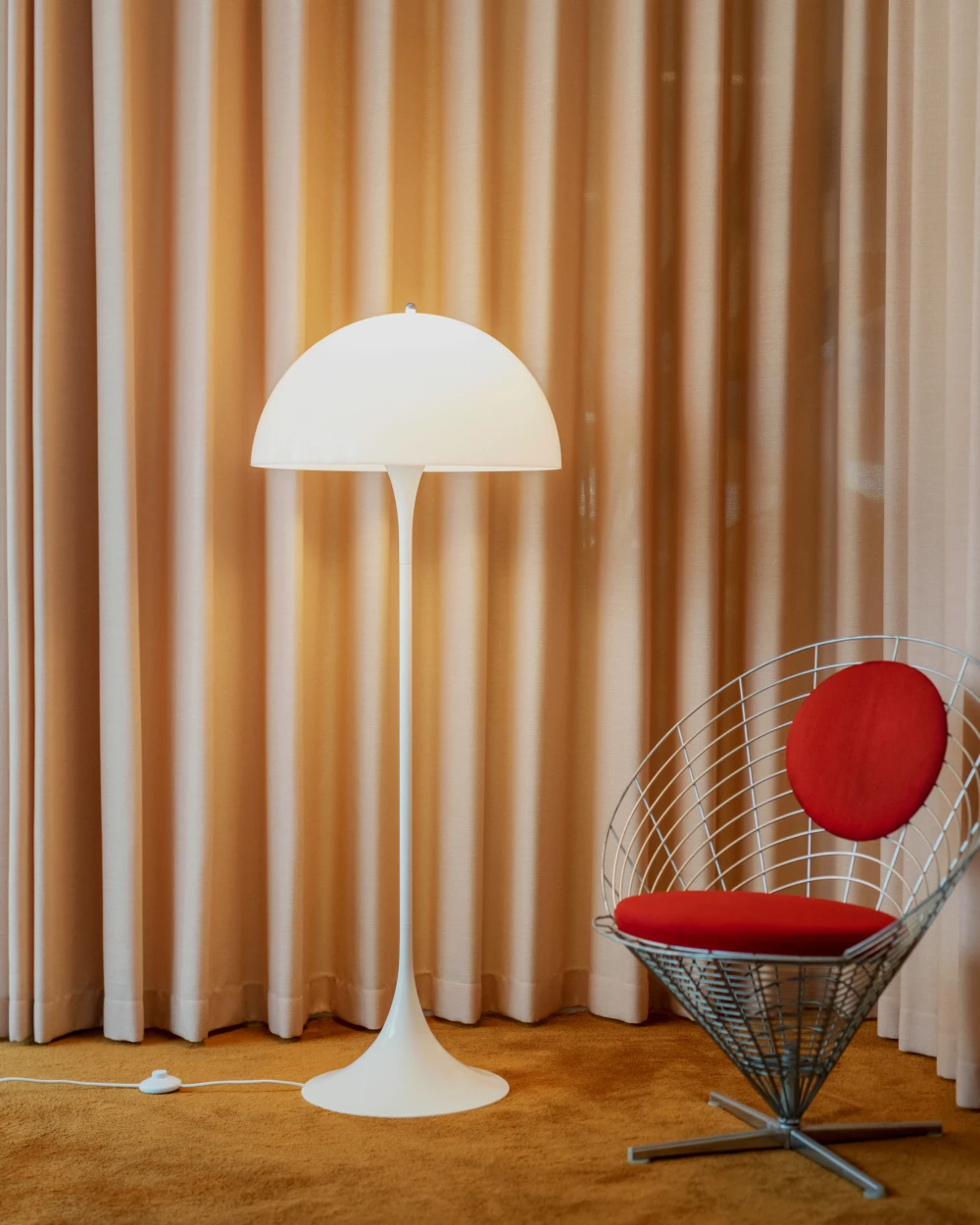 Un lampadaire Panthella blanc se dresse devant des rideaux dorés, près d’un fauteuil en fil métallique aux coussins rouges. © 2026 Louis Poulsen.