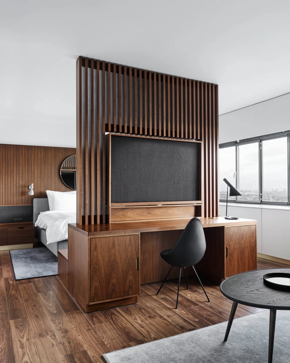 A black AJ Table Lamp and a white AJ Wall Lamp in a hotel room with warm wood panelling and a city view. © 2026 Louis Poulsen.