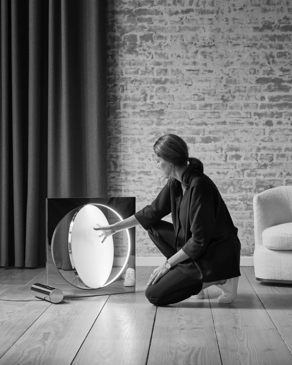 Moonsetter floor lamp with its glowing disc, as a person adjusts it in a room with a brick wall and wood floor. © 2026 Louis Poulsen.
