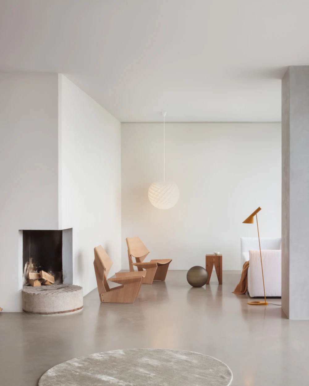 A white Patera Pendant and a brass-toned AJ Floor Lamp in a bright minimalist living room with a fireplace and wooden chairs. © 2026 Louis Poulsen.