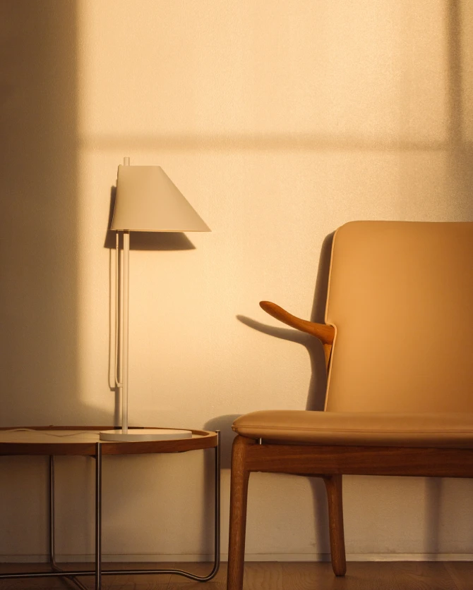 A white AJ Table Lamp stands on a small side table beside a leather chair in warm sunlight. © 2026 Louis Poulsen.