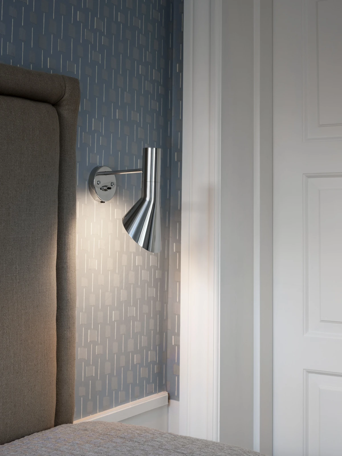 A polished-metal AJ Wall Lamp illuminates the bedside against a blue patterned wall. © 2026 Louis Poulsen.