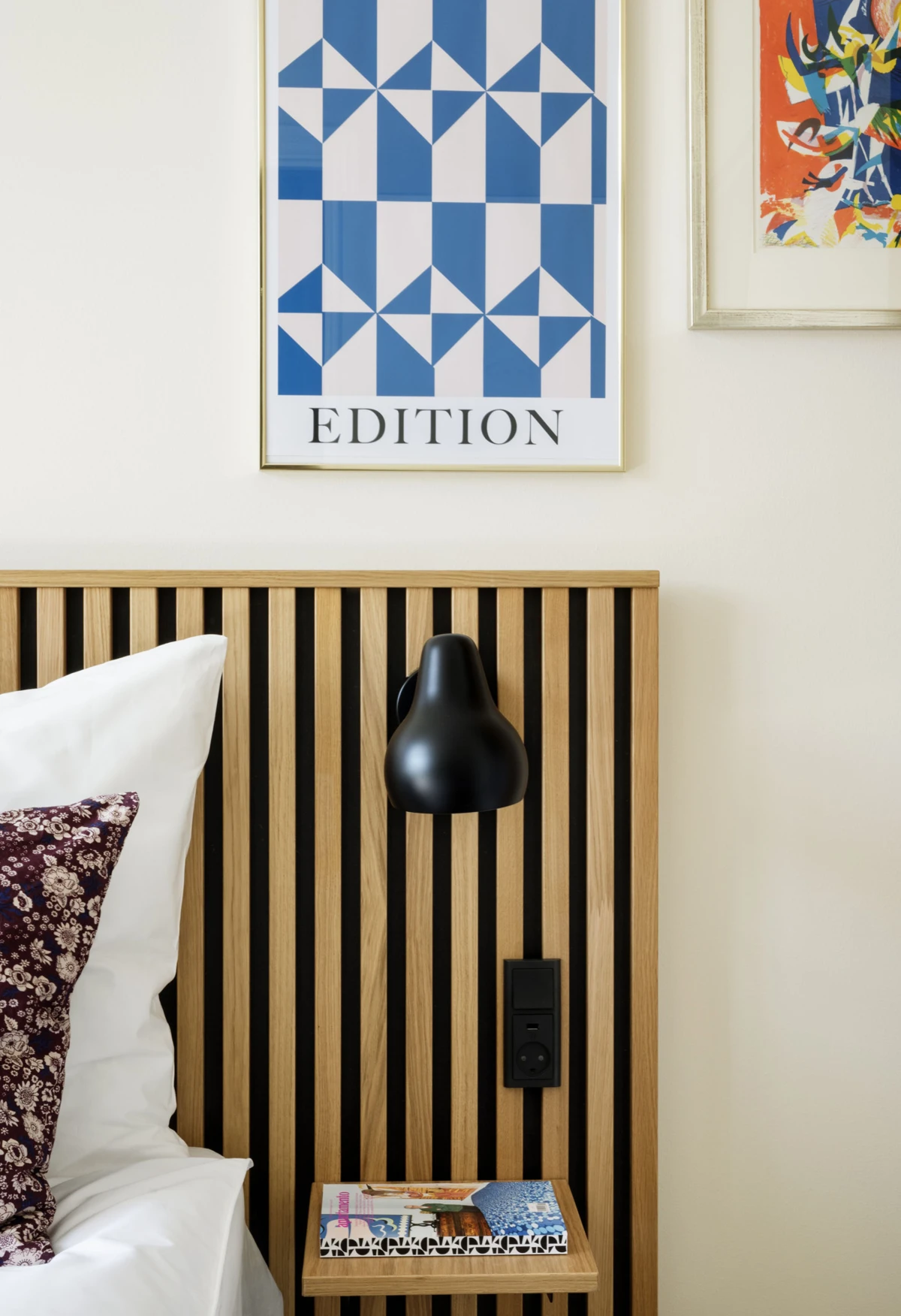 En svart Keglen vägglampa intill en hotellsäng med en sänggavel i ljusa träspjälor. © 2026 Louis Poulsen.
