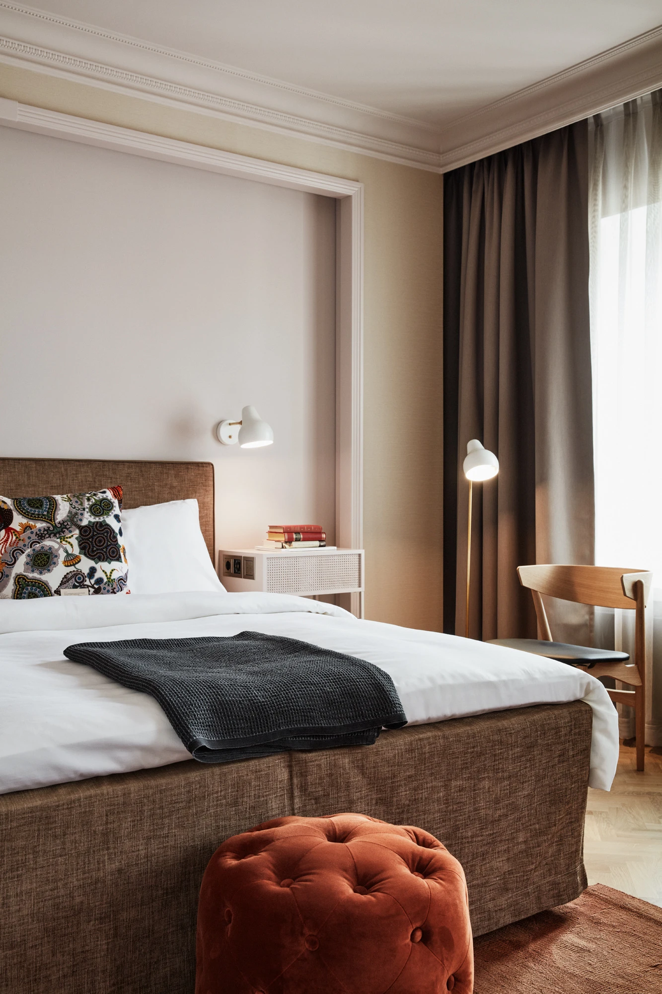 En hvid AJ Wall Lamp og AJ Floor Lamp lyser op i et roligt hotelværelse med seng og mørke gardiner. © 2026 Louis Poulsen.