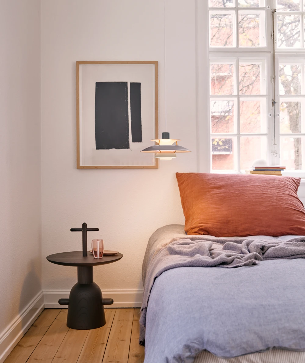 A grey PH 5 pendant hangs beside the bed in a bright bedroom with a timber floor and large windows. © 2026 Louis Poulsen.
