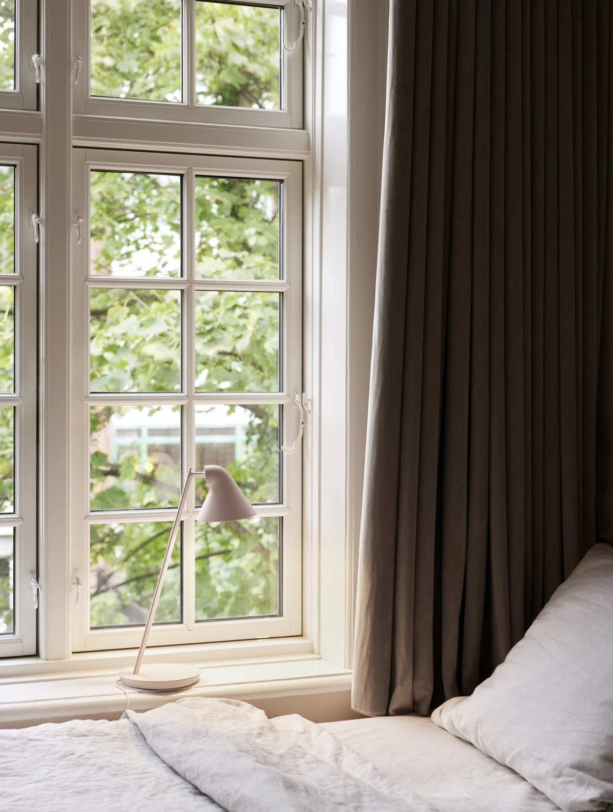 A pale rose NJP Mini Table Lamp sits on the windowsill in a bright bedroom. © 2026 Louis Poulsen.