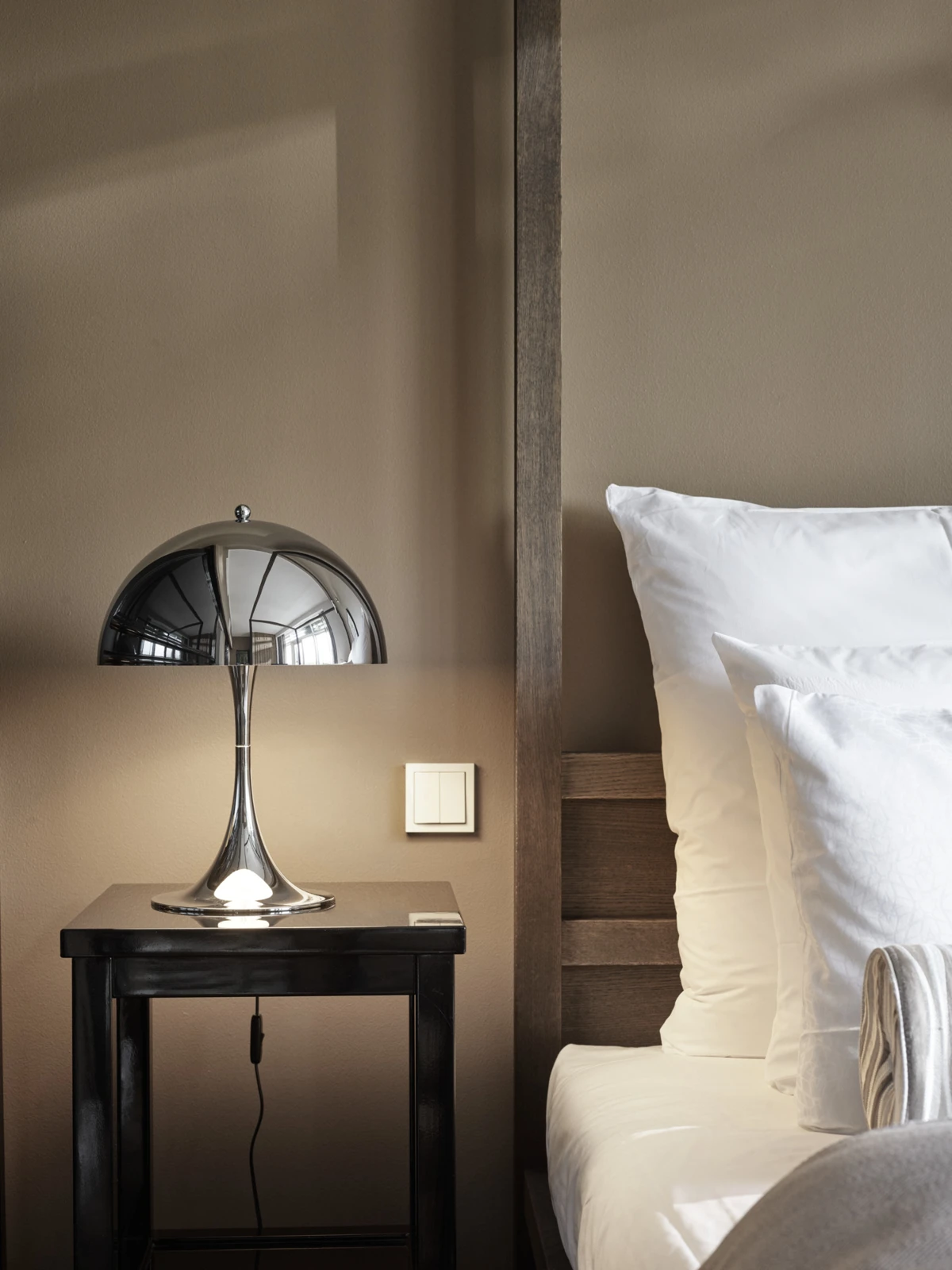 A chrome Panthella 320 Table Lamp sits on a dark bedside table beside a bed with white linens. © 2026 Louis Poulsen.