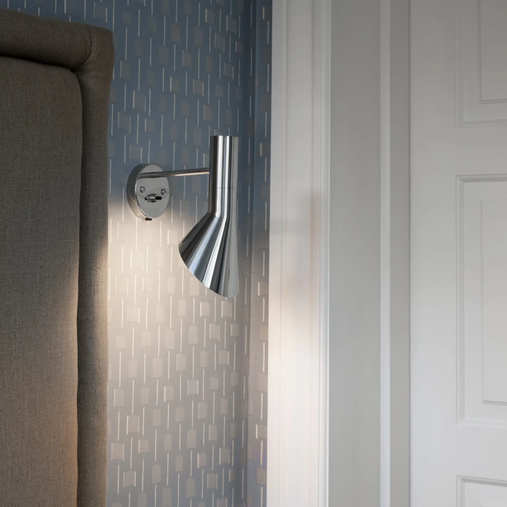 A chrome AJ Wall Lamp beside an upholstered headboard in a bedroom with patterned wallpaper. © 2026 Louis Poulsen.