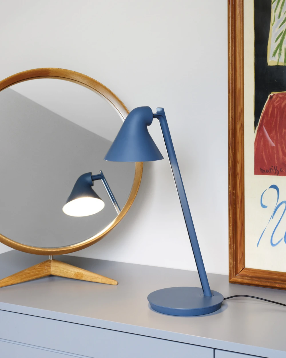 A blue NJP Mini Table Lamp sits on a bedroom dresser beside a round mirror. © 2026 Louis Poulsen.