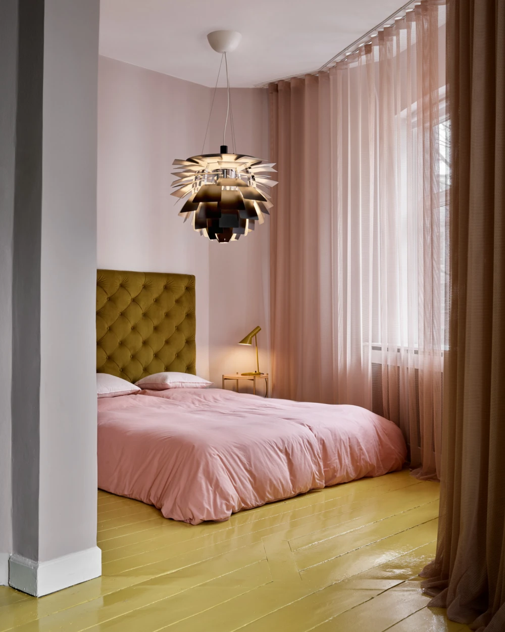 A brass PH Artichoke pendant and an AJ Mini Table Lamp light a bedroom with pink bedding and yellow floorboards. © 2026 Louis Poulsen.