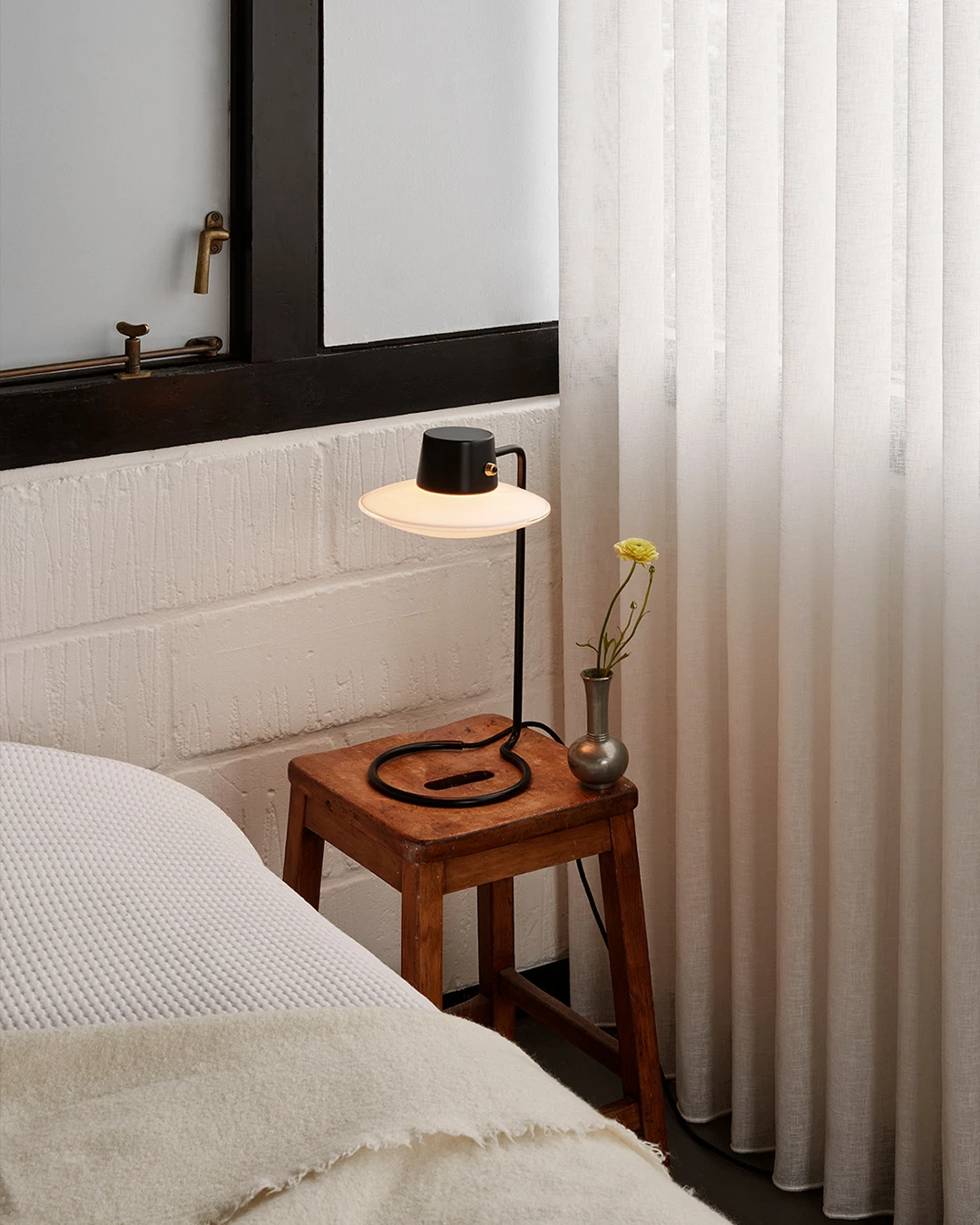 A black AJ Oxford Table Lamp glows on a wooden bedside table beside a bed with white linens. © 2026 Louis Poulsen.