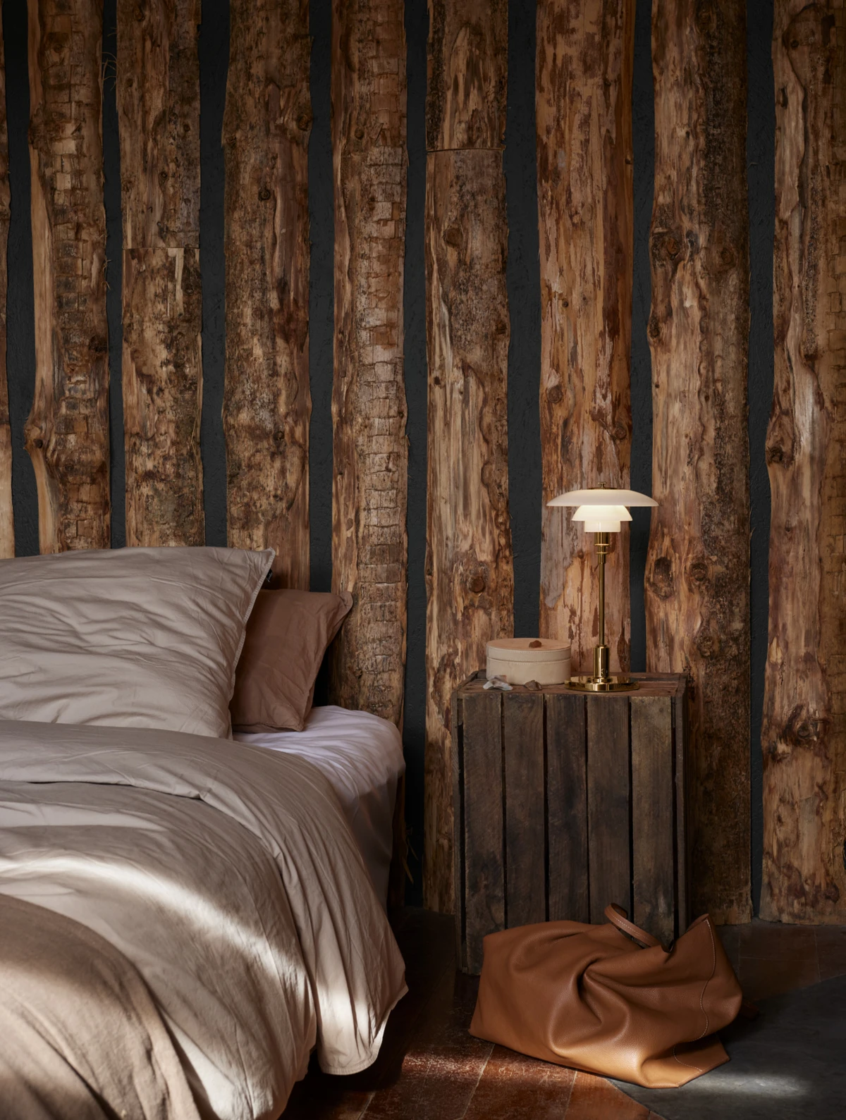 A brass PH 2/1 Portable Lamp glows on a wooden bedside table in a bedroom with rustic timber walls. © 2026 Louis Poulsen.