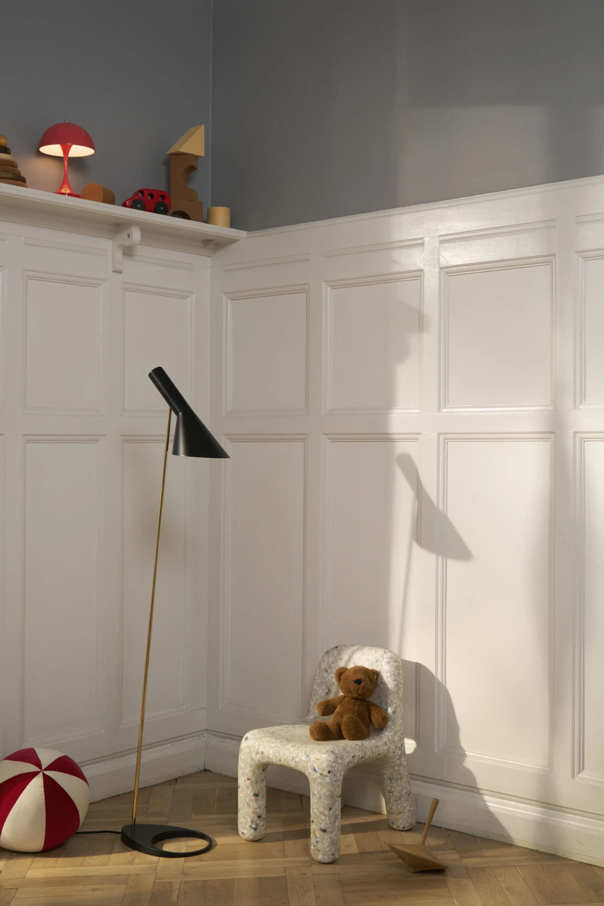 A black AJ Floor Lamp with a brass stem stands in a children’s room beside a small chair and teddy bear. © 2026 Louis Poulsen.