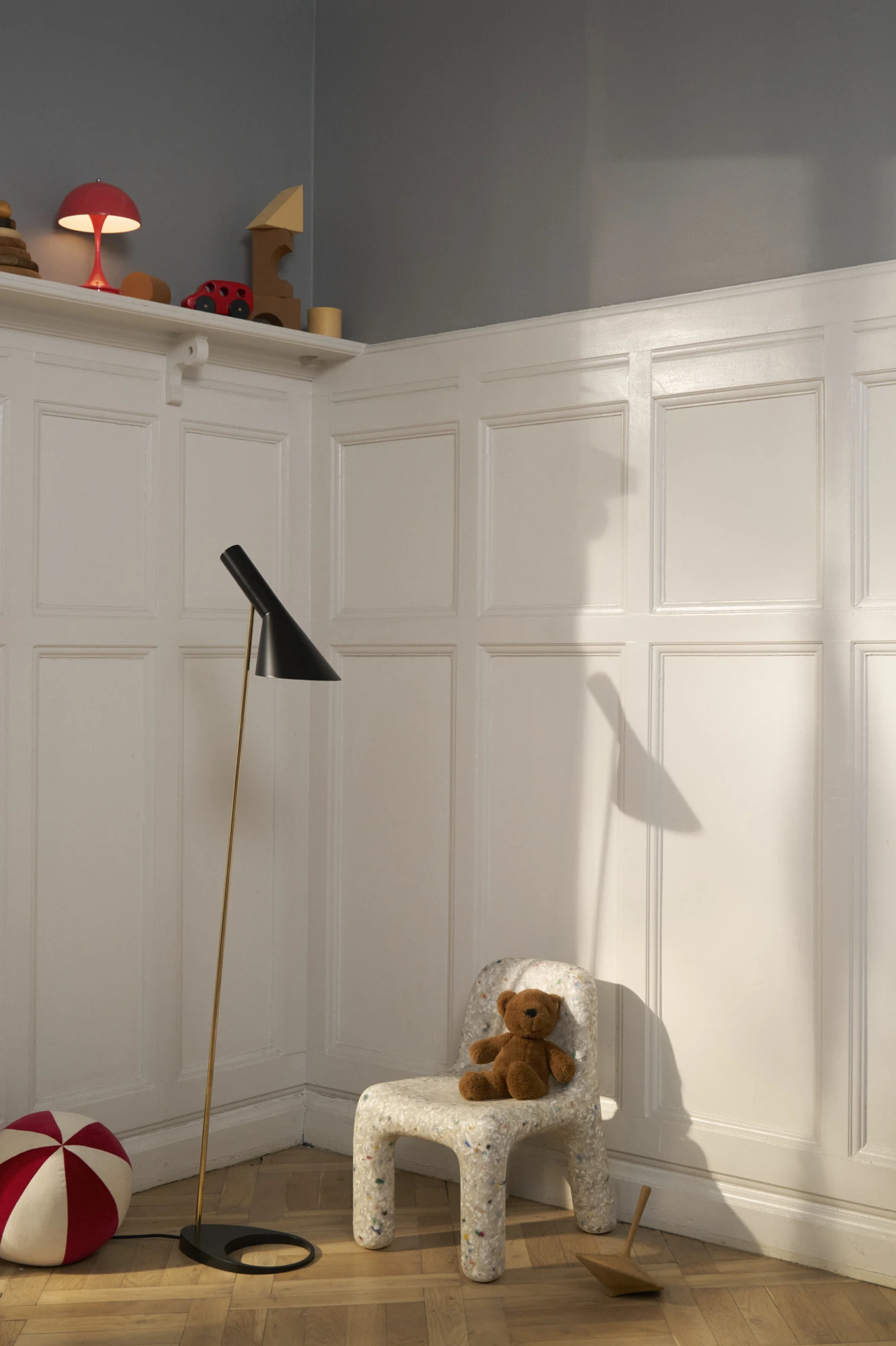 En svart AJ Floor Lamp med mässingsstång står i ett barnrum med vita väggpaneler, en liten stol och en nallebjörn. © 2026 Louis Poulsen.
