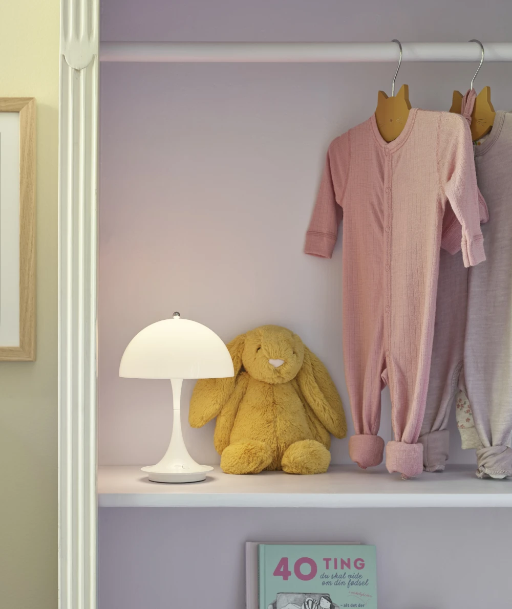 Een witte Panthella 250 tafellamp staat naast een knuffelkonijn en babykleding op een plank in een kinderkamer. © 2026 Louis Poulsen.