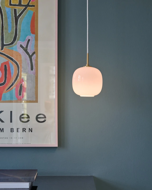A VL 45 Radiohus Pale Rose pendant glows above a desk against a blue-green wall beside colourful artwork. © 2026 Louis Poulsen.