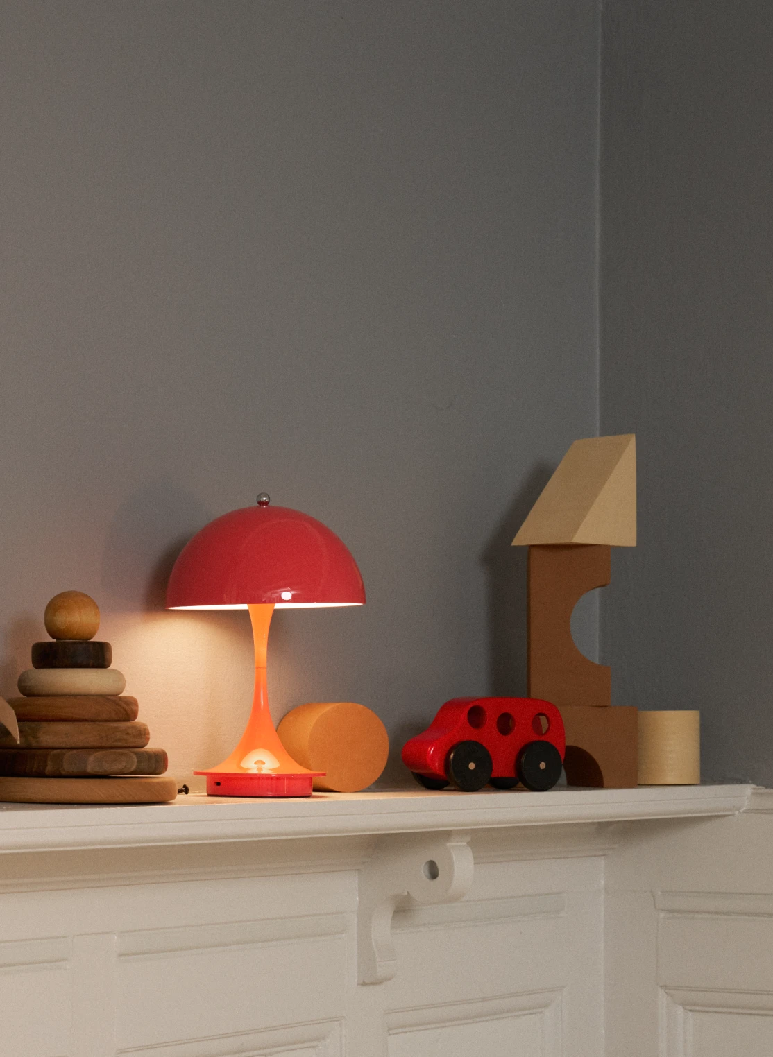 A red and orange Panthella 160 Portable Lamp glows on a shelf among toys in a children’s room. © 2026 Louis Poulsen.