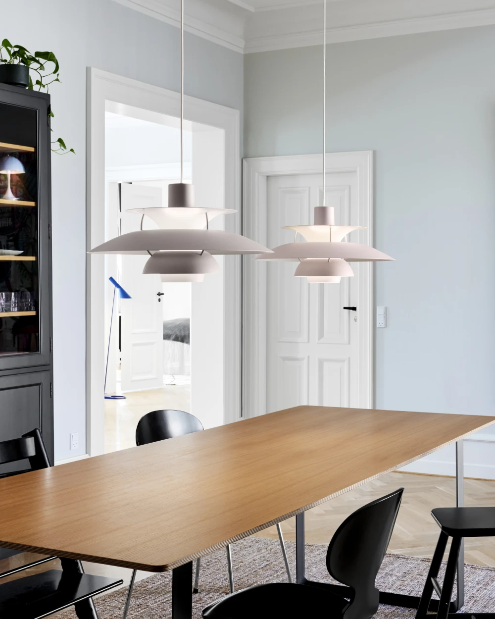 Two light-coloured PH 5 pendants hang over a wooden dining table in a bright room with black chairs. © 2026 Louis Poulsen.