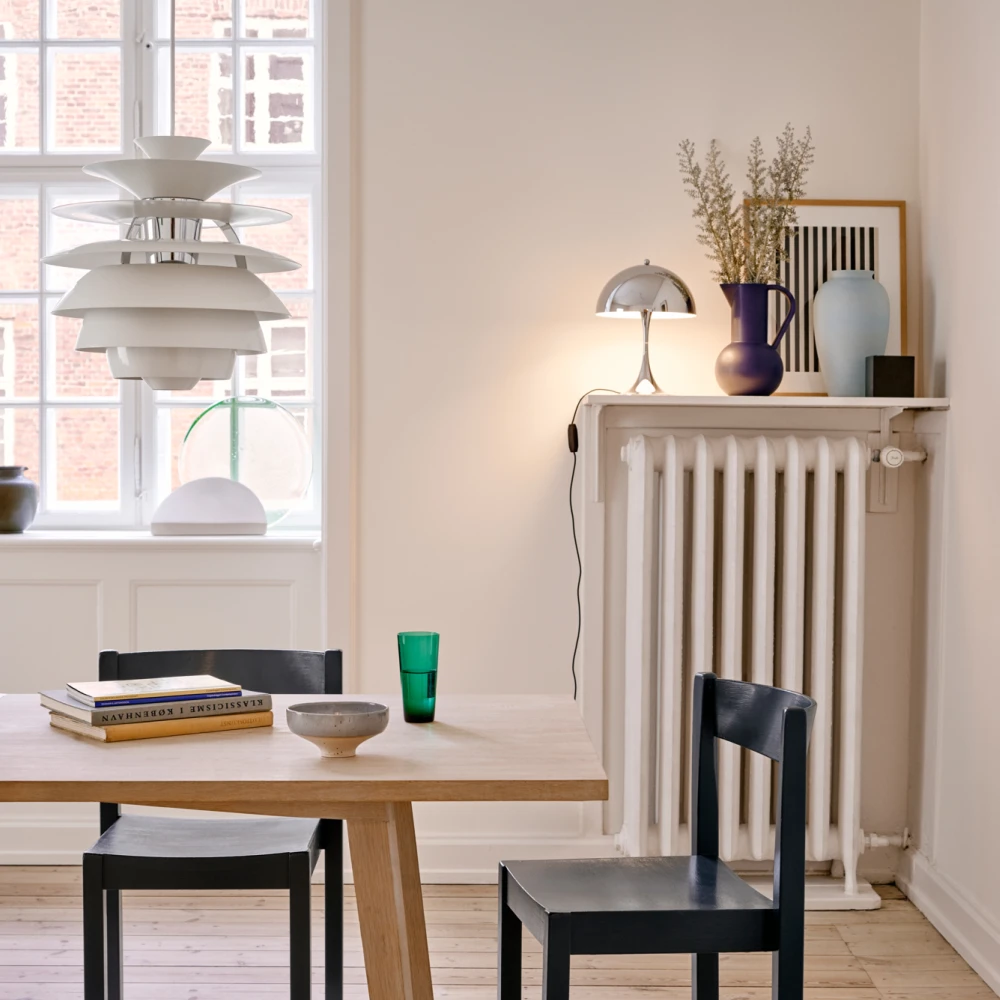 A white PH Snowball pendant above a dining table in a bright room with black chairs and a plank floor. © 2026 Louis Poulsen.