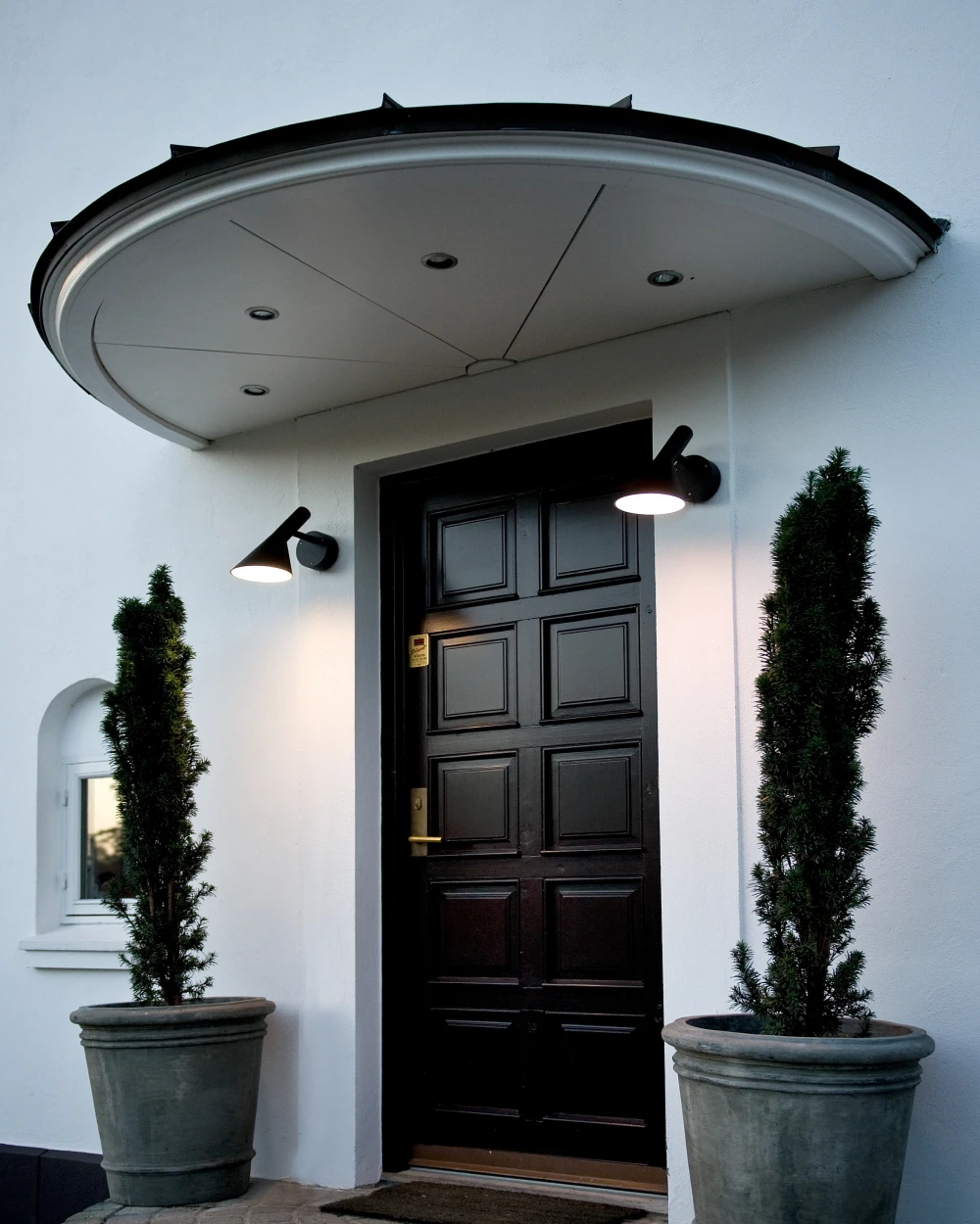 Two black AJ Wall Lamp wall lights illuminate the entrance of a white house with a black door and potted evergreens. © 2026 Louis Poulsen.