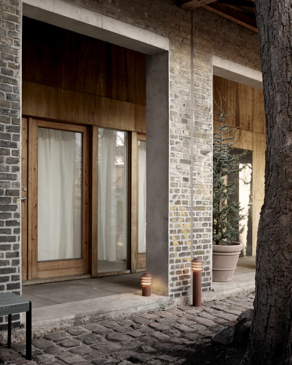 Two terracotta Bysted Garden Bollards stand beside a brick-and-wood entrance and a cobbled path. © 2026 Louis Poulsen.