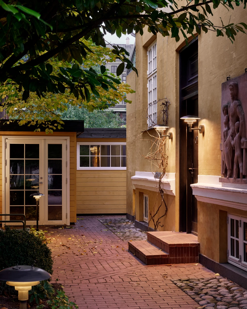 PH 3-2½ Outdoor Wall Lamps on a yellow facade and PH 3-2½ Bollards lighting a paved entrance courtyard. © 2026 Louis Poulsen.