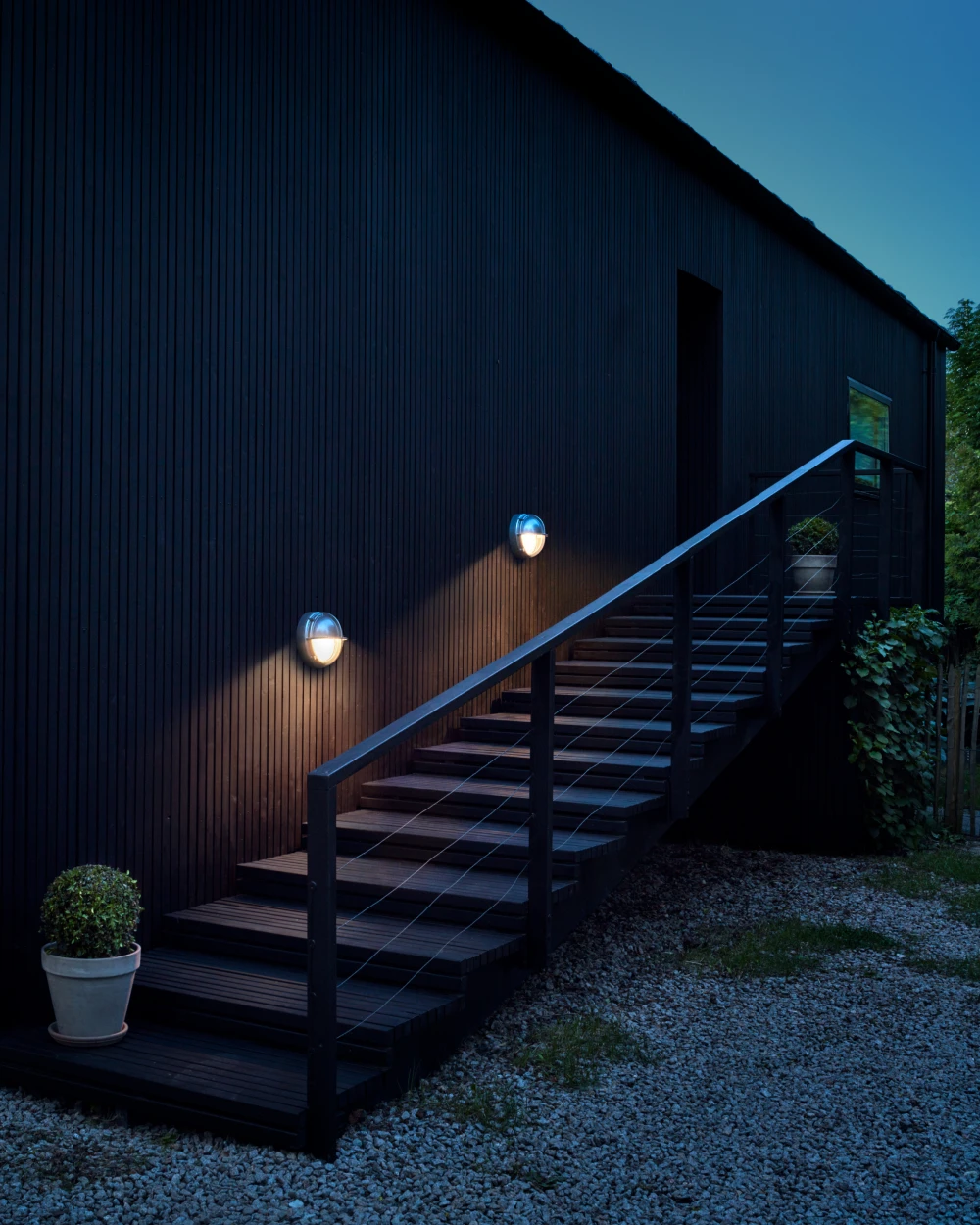 Two illuminated Skot Wall Lamps on a dark timber facade beside an outdoor entrance staircase. © 2026 Louis Poulsen.