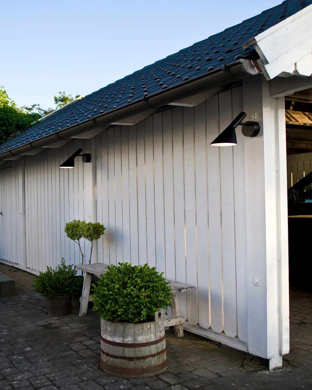 Two black AJ Wall Lamp wall lights on the white wooden exterior of a garage beneath a dark tiled roof. © 2026 Louis Poulsen.