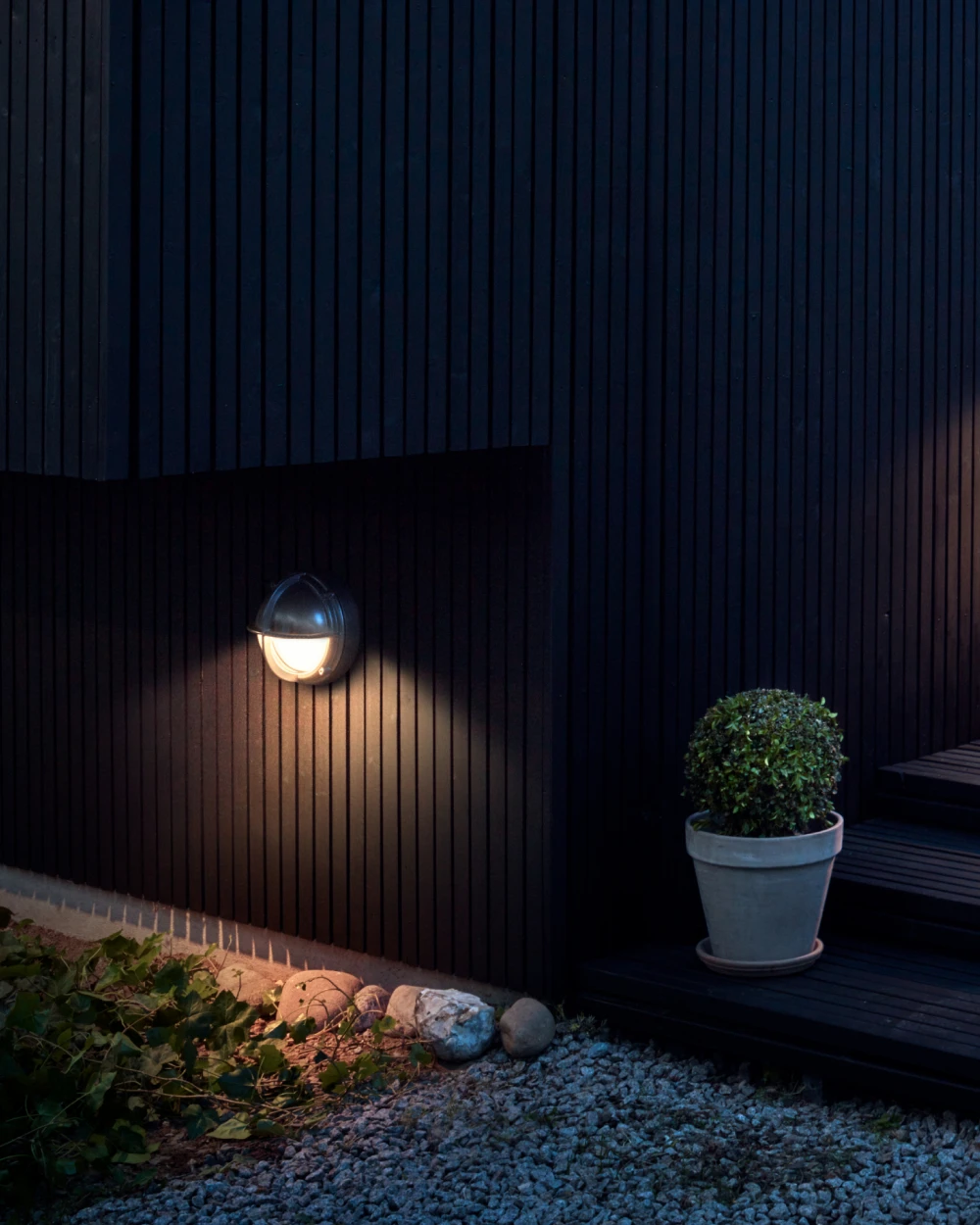 A Skot Wall Lamp casts a warm glow on dark vertical cladding beside a gravel path and potted shrub. © 2026 Louis Poulsen.