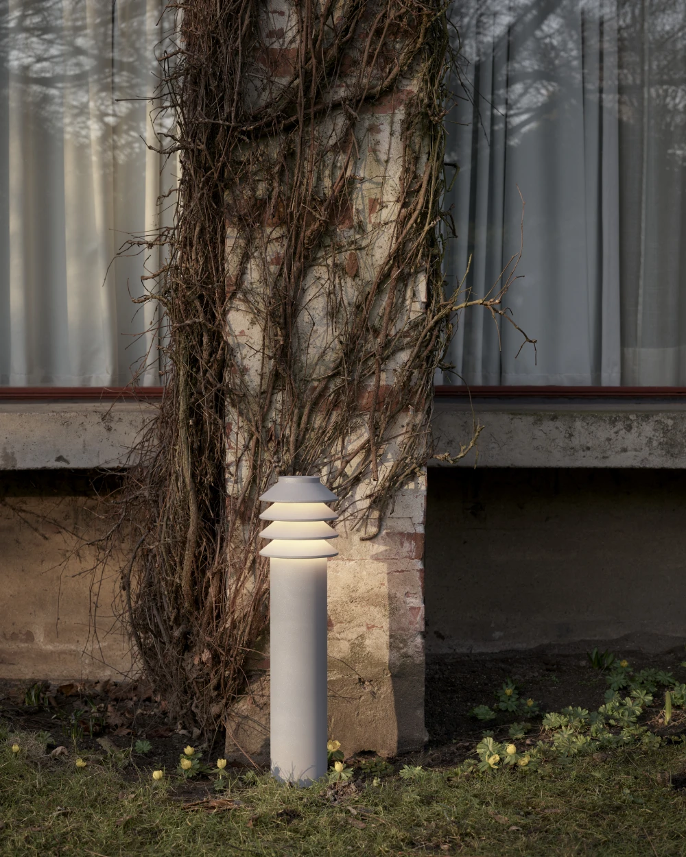 A grey Bysted Garden Bollard glows beside a vine-covered wall in a garden. © 2026 Louis Poulsen.