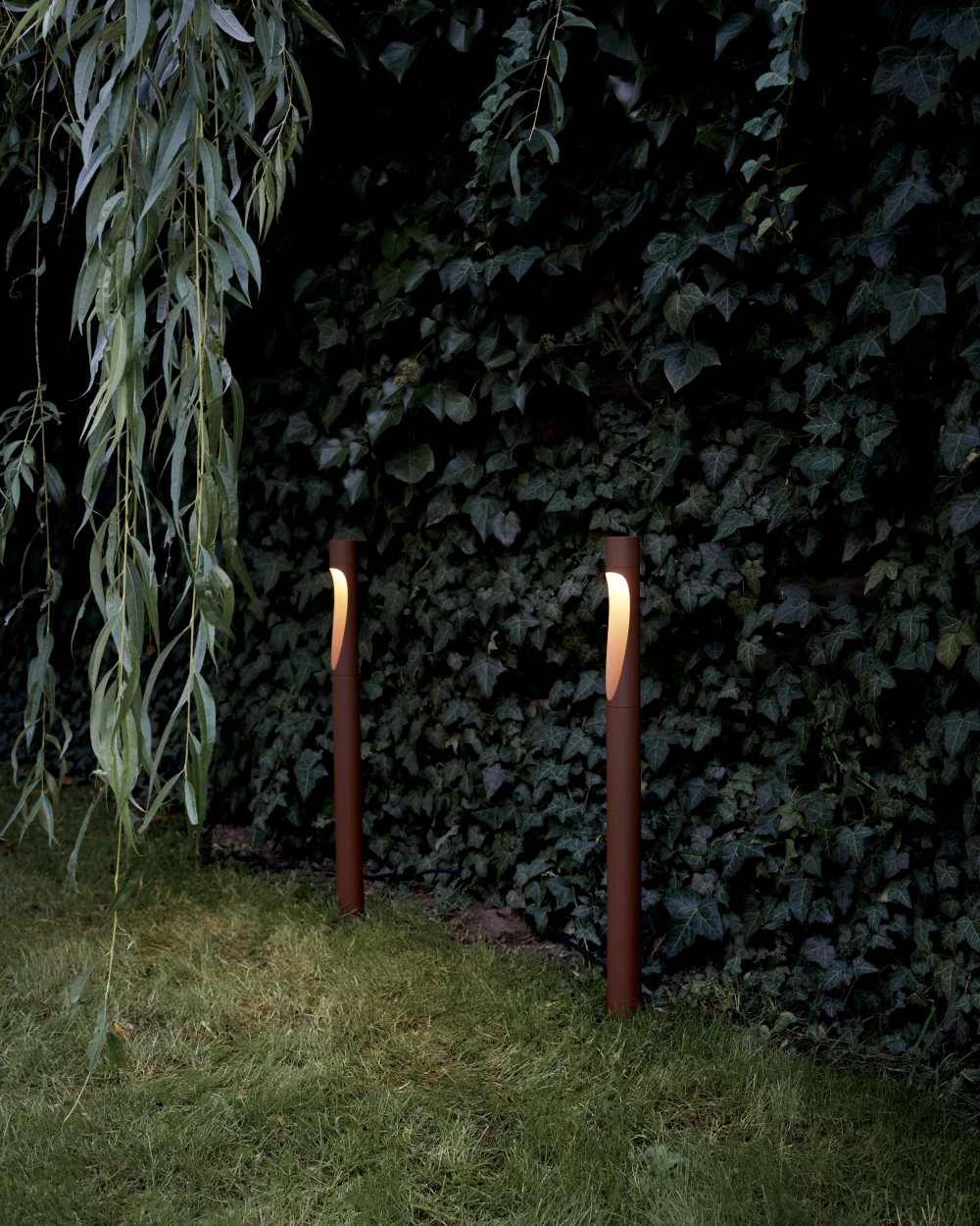 Two brown Flindt Garden Bollards illuminate a grass path beside a dense, leafy hedge. © 2026 Louis Poulsen.